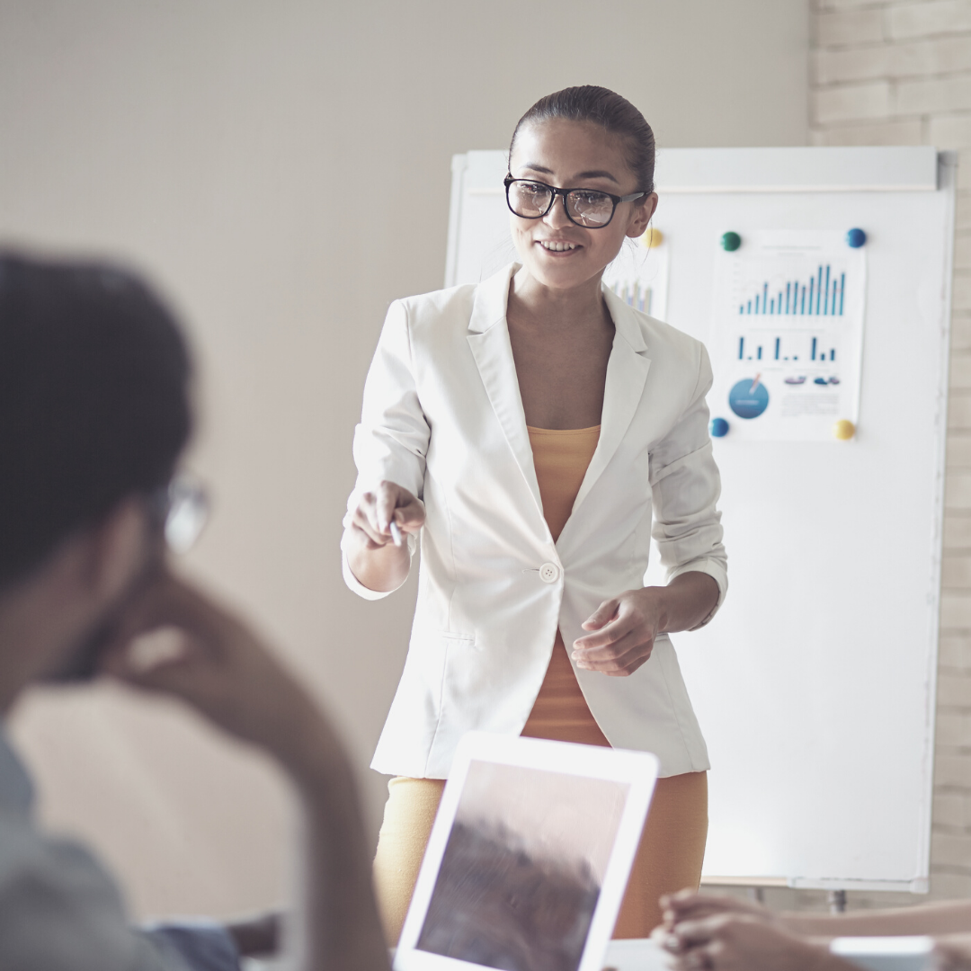 female manager wearing orange dress and white blazer pointing and standing in front of flip chart